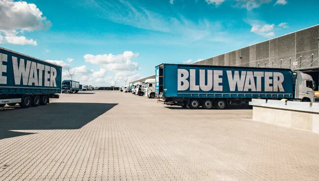 Kontainer DAM_Blue Water Case_Trucks Blue Water trucks outside a warehouse under a blue sky.